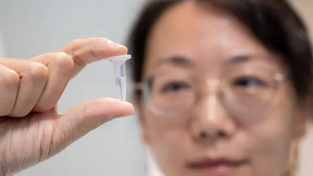 Close-up of a researcher's fingers holding a small vial of light-sensitive nanoparticle solution up to the light, with Aarhus University associate professor Menglin Chen looking at it in the blurred background.