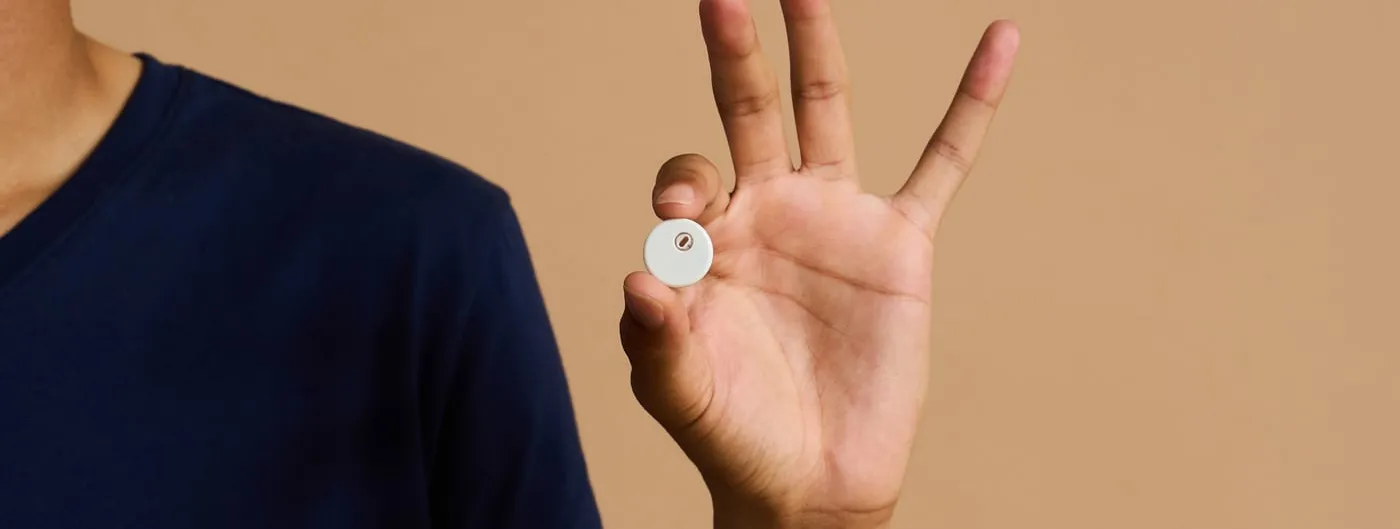 A person in a navy T-shirt holds a small white circular sensor disc between thumb and finger against a beige backdrop.
