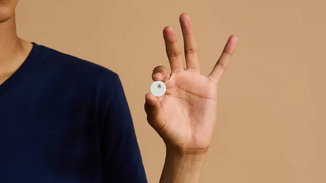 A person in a navy T-shirt holds a small white circular sensor disc between thumb and finger against a beige backdrop.