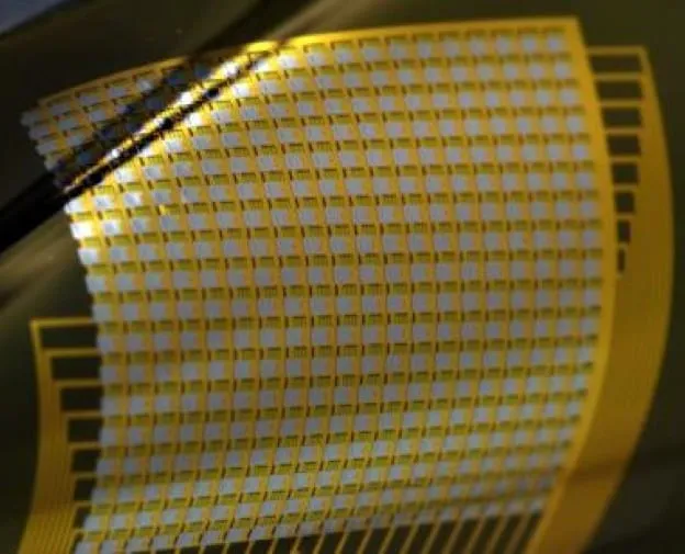A thin, flexible microelectrode array on a transparent polymer film, its grid of gold contacts and traces catching the light as the sheet curls away from a dark surface.