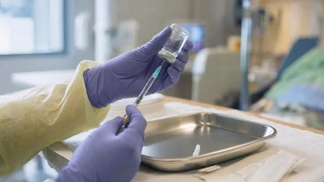 Close-up photograph of a gloved clinician's hands holding a small glass medication vial and a syringe above a stainless steel tray in a hospital treatment room.