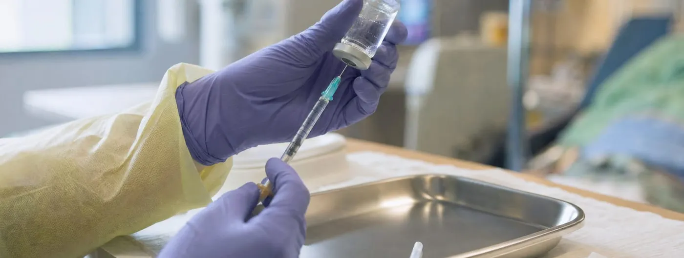 Close-up photograph of a gloved clinician's hands holding a small glass medication vial and a syringe above a stainless steel tray in a hospital treatment room.