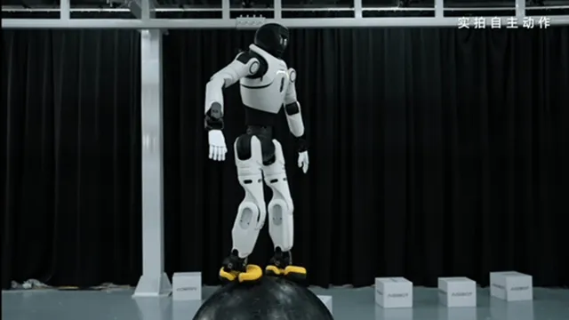 The white and black Lingxi X2 humanoid robot stands balanced on top of a large black ball in a studio, arms held out to the sides, with branded boxes stacked on the floor behind it.