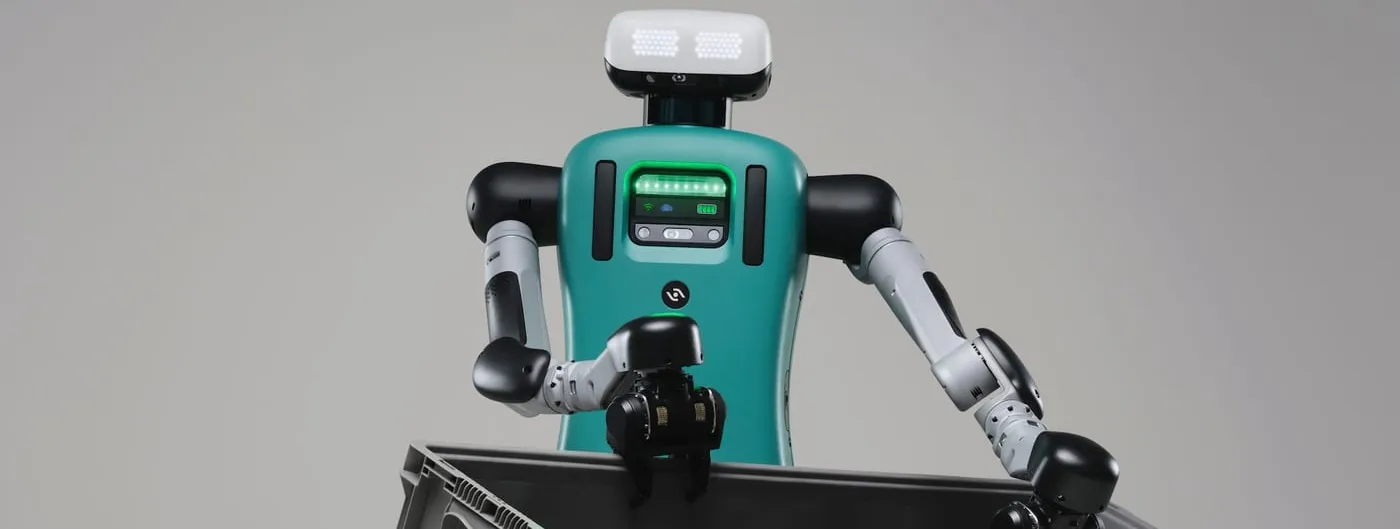 A studio photograph of a teal-and-grey humanoid robot with a rectangular LED face, holding a large grey plastic tote out in front of it with two pincer hands.