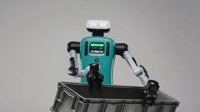 A studio photograph of a teal-and-grey humanoid robot with a rectangular LED face, holding a large grey plastic tote out in front of it with two pincer hands.
