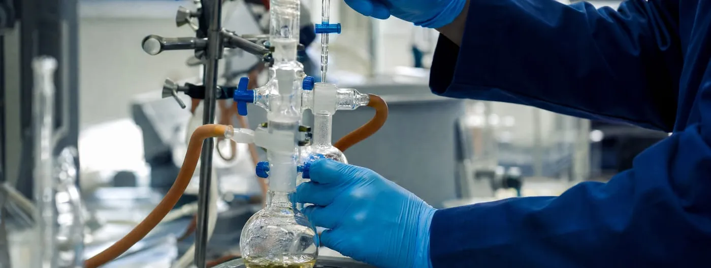 Close-up photograph of a gloved researcher's hands adjusting glass laboratory apparatus and a pipette above a round-bottom flask during small-molecule drug synthesis work.