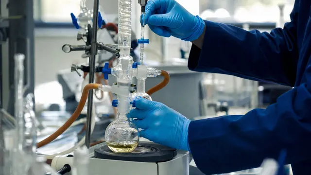 Close-up photograph of a gloved researcher's hands adjusting glass laboratory apparatus and a pipette above a round-bottom flask during small-molecule drug synthesis work.