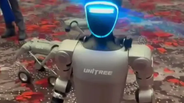 A knee-high humanoid robot with a glowing blue visor walks across the patterned carpet of a convention hall while people stand in the background.