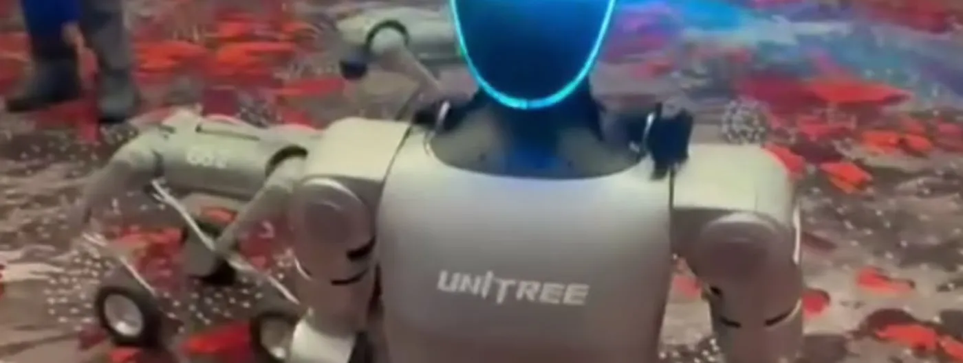 A knee-high humanoid robot with a glowing blue visor walks across the patterned carpet of a convention hall while people stand in the background.