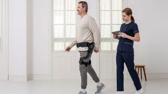 A man walks across a bright white room wearing the Angel Suit H10 — a powered brace strapped at the waist and along one thigh — while a clinician in navy scrubs follows him holding a tablet.