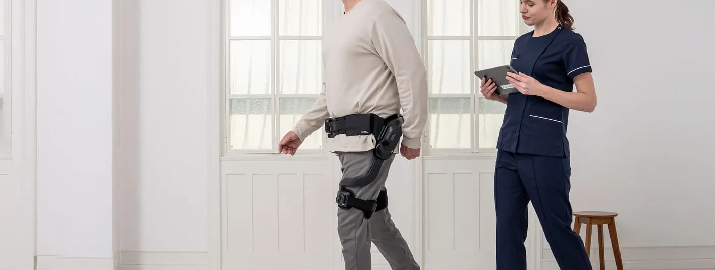 A man walks across a bright white room wearing the Angel Suit H10 — a powered brace strapped at the waist and along one thigh — while a clinician in navy scrubs follows him holding a tablet.
