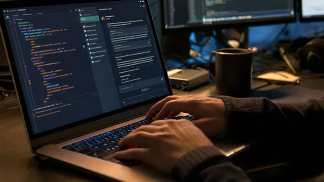 A software engineer's hands type on a laptop keyboard showing a code editor and chat panel, with two more monitors displaying code in a dim night-time office.