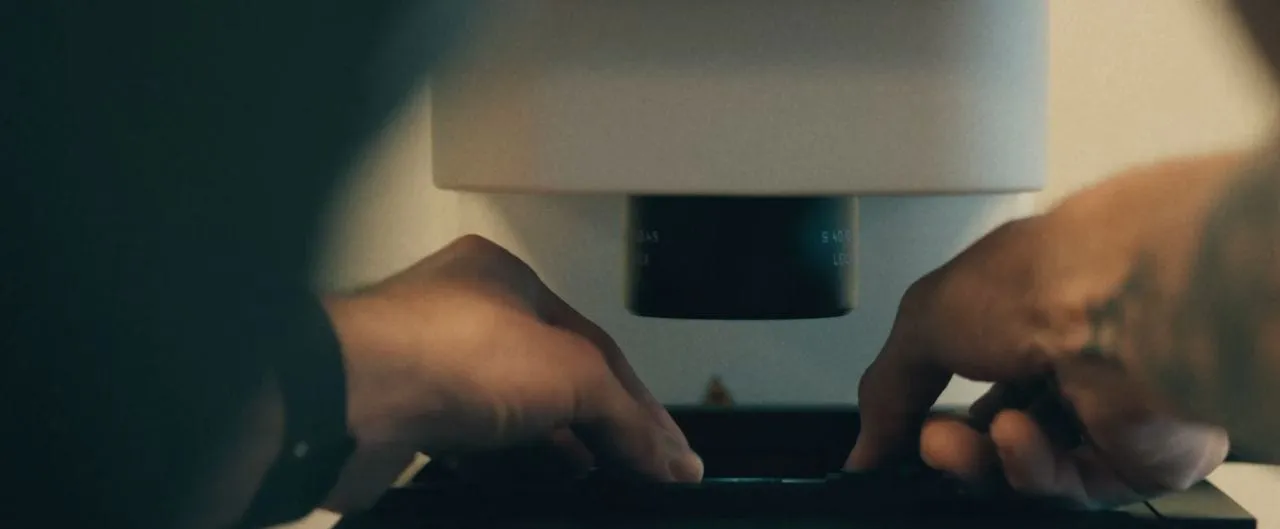 Two hands adjust a specimen stage beneath the objective lens of a laboratory microscope in dim light, the instrument housing pale against a dark background.