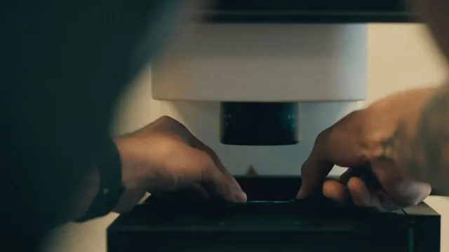 Two hands adjust a specimen stage beneath the objective lens of a laboratory microscope in dim light, the instrument housing pale against a dark background.