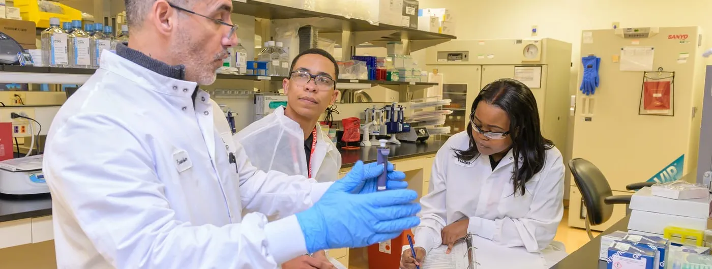 Three researchers in white coats examine a small vial in a laboratory, with pipettes, reagent boxes and racks of sample tubes on the benches around them.