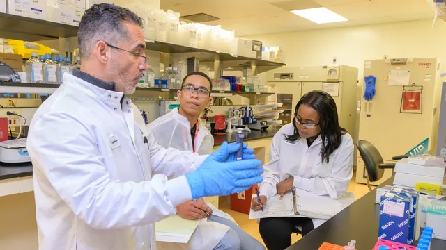 Three researchers in white coats examine a small vial in a laboratory, with pipettes, reagent boxes and racks of sample tubes on the benches around them.