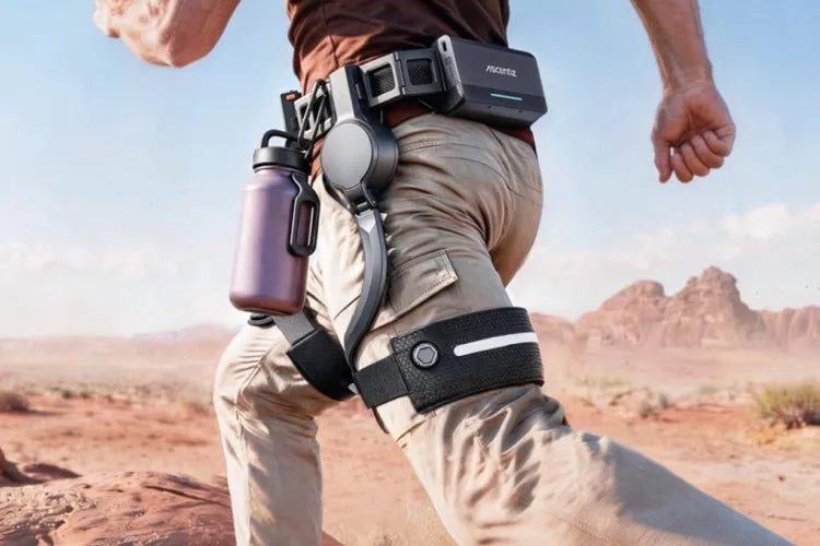 A runner in tan trousers crossing red desert ground, wearing a black powered belt module at the hip with a rigid arm running down the outside of the thigh to a strap above the knee.
