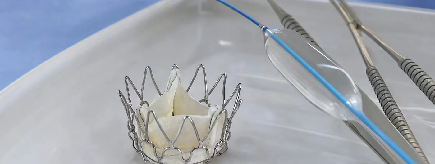 A generic expandable prosthetic heart valve with a thin metal wire frame and pale leaflets sits on a sterile tray in an operating room, beside a balloon catheter and two surgical forceps.