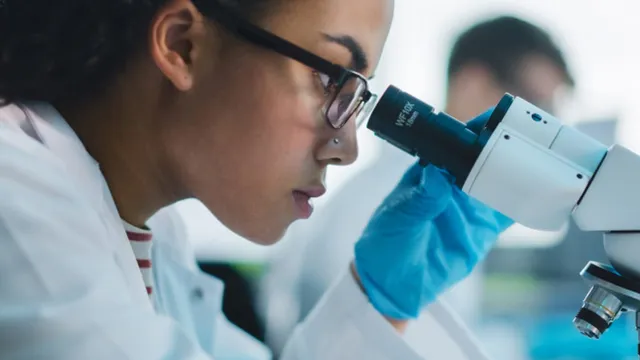 A researcher wearing glasses and a blue nitrile glove peers into a laboratory microscope's eyepiece while examining a specimen.