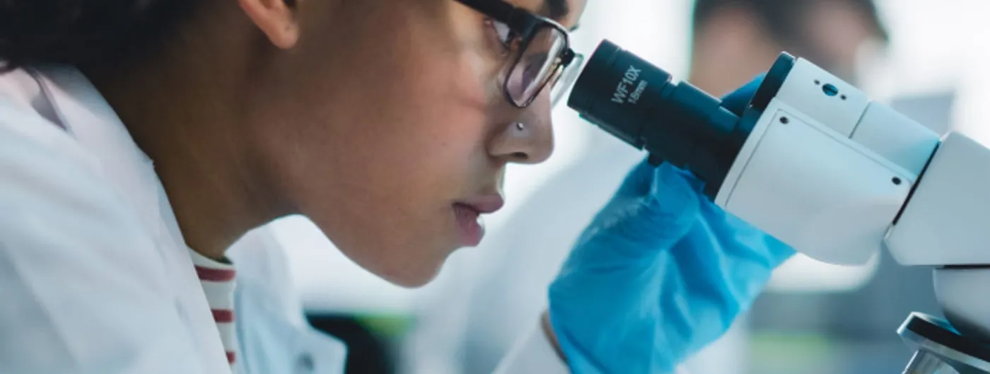 A researcher wearing glasses and a blue nitrile glove peers into a laboratory microscope's eyepiece while examining a specimen.