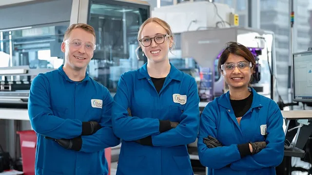 Three Beam Therapeutics scientists in blue lab coats, safety glasses and name badges stand with arms crossed in a laboratory in front of automated lab equipment and a monitor.