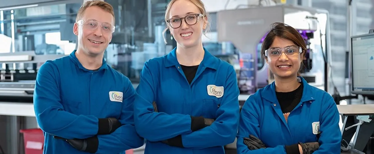 Three Beam Therapeutics scientists in blue lab coats, safety glasses and name badges stand with arms crossed in a laboratory in front of automated lab equipment and a monitor.