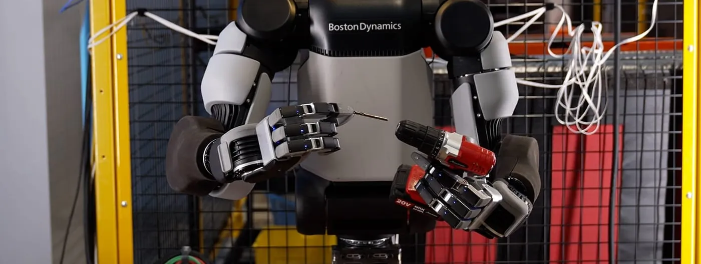 An Atlas humanoid robot with two new four-fingered hands holds a small drill bit in its left hand and a red cordless drill in its right, standing at a workbench inside a yellow safety cage.