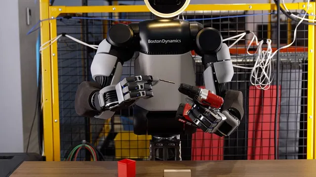 An Atlas humanoid robot with two new four-fingered hands holds a small drill bit in its left hand and a red cordless drill in its right, standing at a workbench inside a yellow safety cage.