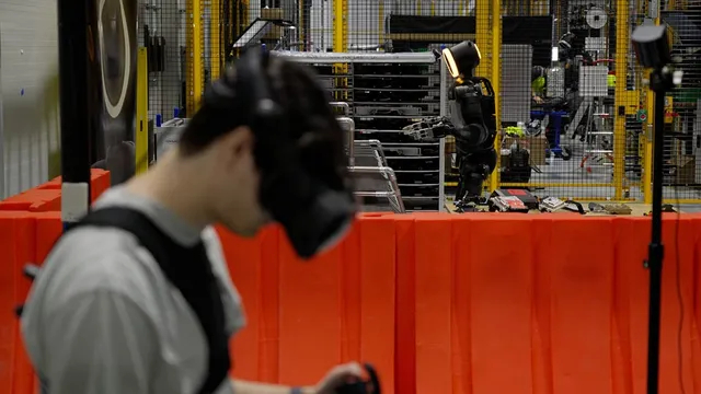 A trainer wearing a VR headset and haptic controllers teleoperates a Boston Dynamics Atlas humanoid robot as it reaches toward a parts rack inside the Robotics Metaplant Application Center, with factory workers visible behind a safety fence.