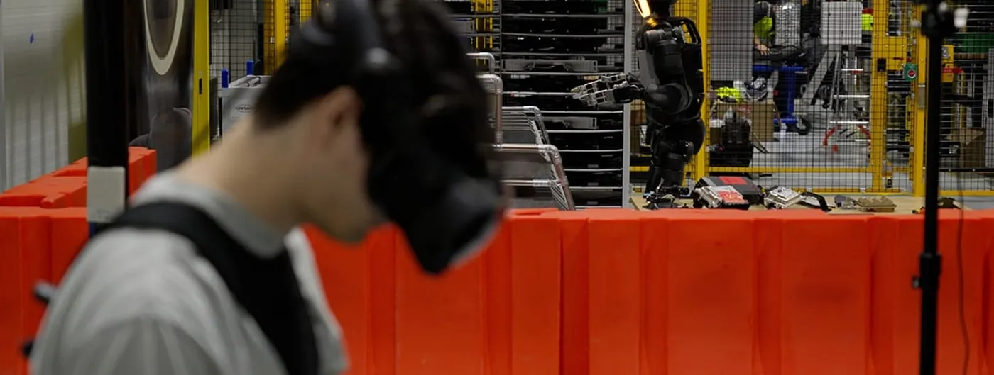 A trainer wearing a VR headset and haptic controllers teleoperates a Boston Dynamics Atlas humanoid robot as it reaches toward a parts rack inside the Robotics Metaplant Application Center, with factory workers visible behind a safety fence.
