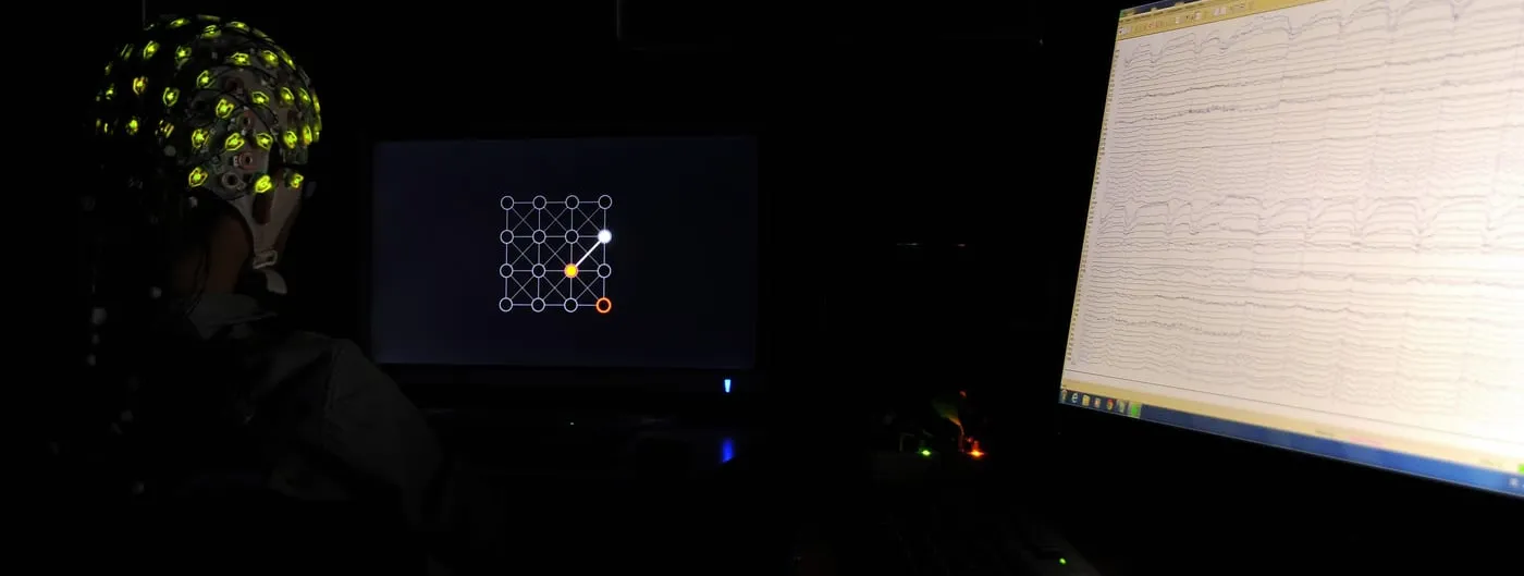 A research participant wearing a non-invasive EEG electrode cap, seen from behind, facing a display showing a brain-computer interface grid task, with a second monitor streaming live EEG traces.