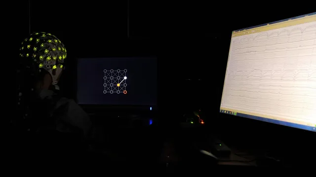 A research participant wearing a non-invasive EEG electrode cap, seen from behind, facing a display showing a brain-computer interface grid task, with a second monitor streaming live EEG traces.