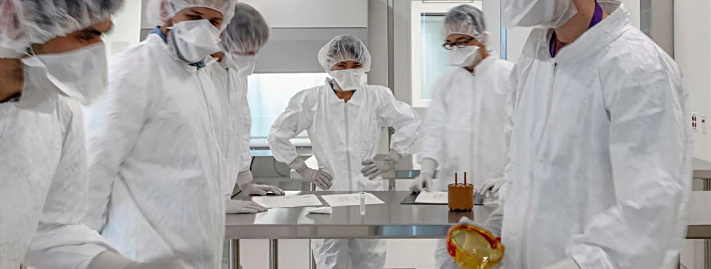 Cleanroom technicians in white PPE gowns, hairnets, masks and gloves work around a steel bench handling small cell-therapy vials and containers inside a CAR T-cell manufacturing facility.