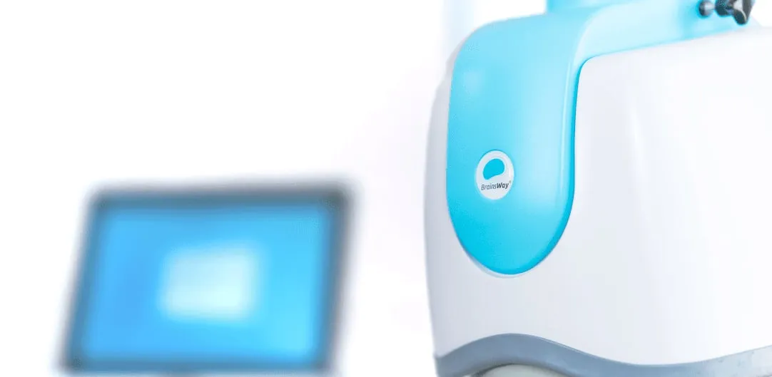 A white and pale blue transcranial magnetic stimulation helmet unit on its positioning arm, with a blurred treatment screen beside it.
