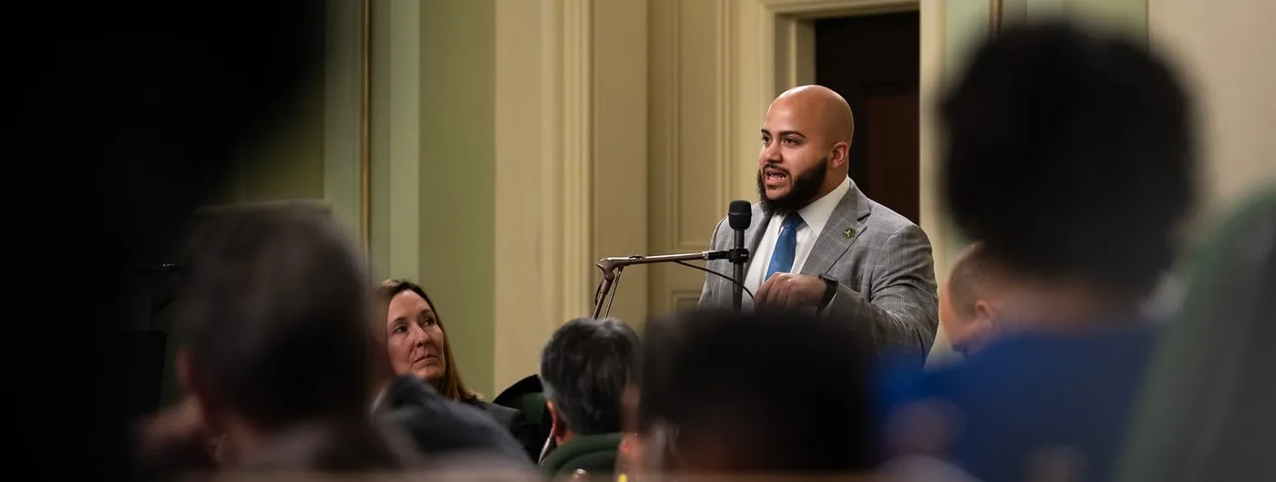 Assemblymember Isaac Bryan speaks into a microphone on the California Assembly floor, seen past the blurred heads of seated colleagues.