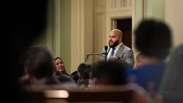 Assemblymember Isaac Bryan speaks into a microphone on the California Assembly floor, seen past the blurred heads of seated colleagues.