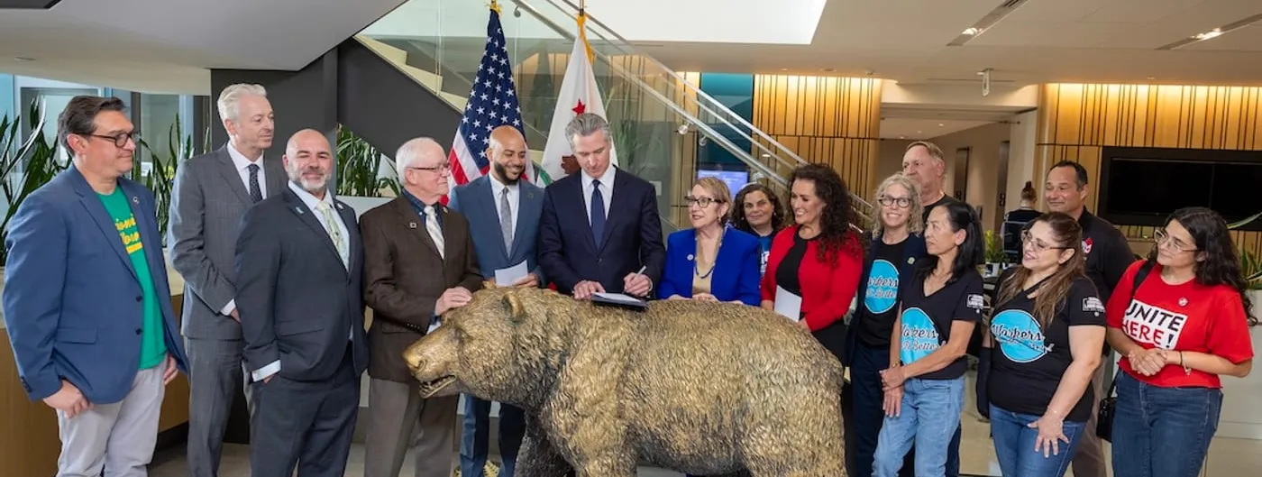 Governor Gavin Newsom signing worker-protection legislation at a table behind a bronze California grizzly bear statue, flanked by legislators and labor union members in a modern atrium with the US and California flags behind him.