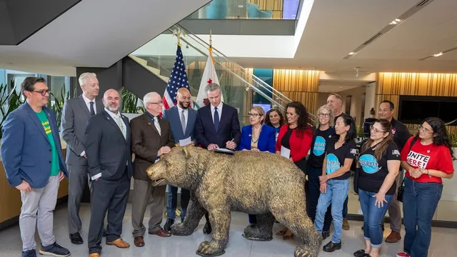 Governor Gavin Newsom signing worker-protection legislation at a table behind a bronze California grizzly bear statue, flanked by legislators and labor union members in a modern atrium with the US and California flags behind him.