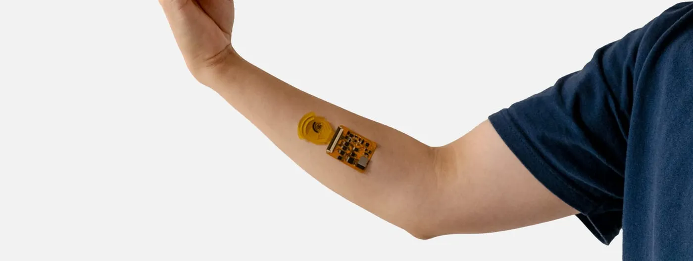 A bare forearm with a small orange circuit board and yellow sensor strip stuck to the inner arm, the hand above it holding a burger.