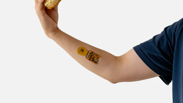 A bare forearm with a small orange circuit board and yellow sensor strip stuck to the inner arm, the hand above it holding a burger.