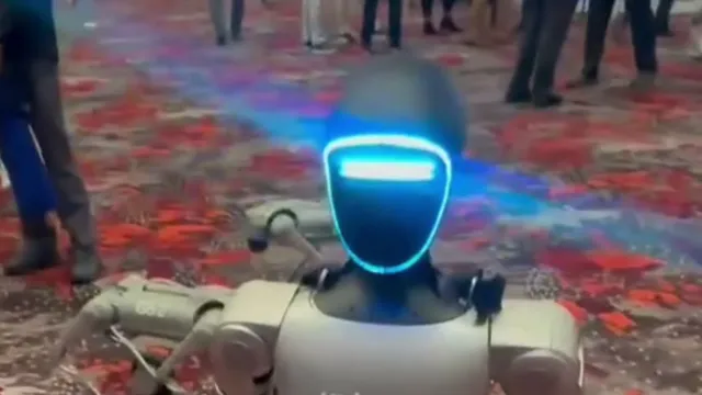 A Unitree humanoid robot with a glowing blue visor-style head and grey exoskeleton frame walks across a patterned carpet at a trade expo, with legs of attendees visible in the background.