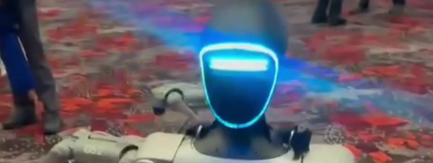 A Unitree humanoid robot with a glowing blue visor-style head and grey exoskeleton frame walks across a patterned carpet at a trade expo, with legs of attendees visible in the background.