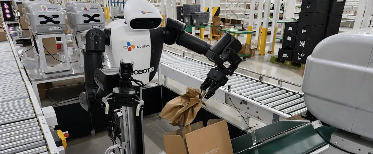 A white and black dual-arm humanoid robot on a wheeled column stands at a roller conveyor in a warehouse, one gripper holding a crumpled sheet of brown packing paper above an open cardboard box, with rows of shelving and stacked crates behind it.