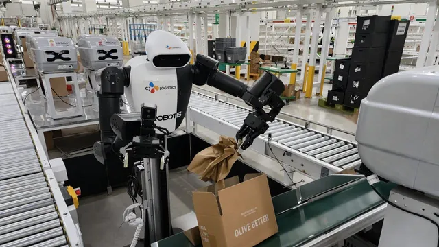 A white and black dual-arm humanoid robot on a wheeled column stands at a roller conveyor in a warehouse, one gripper holding a crumpled sheet of brown packing paper above an open cardboard box, with rows of shelving and stacked crates behind it.
