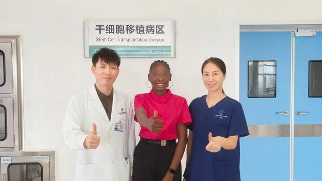 A doctor in a white coat, a patient in a pink shirt whose face is blurred for privacy, and a nurse in navy scrubs stand together giving thumbs up in a hospital corridor beneath a sign reading "Stem Cell Transplantation Division" in Chinese and English.