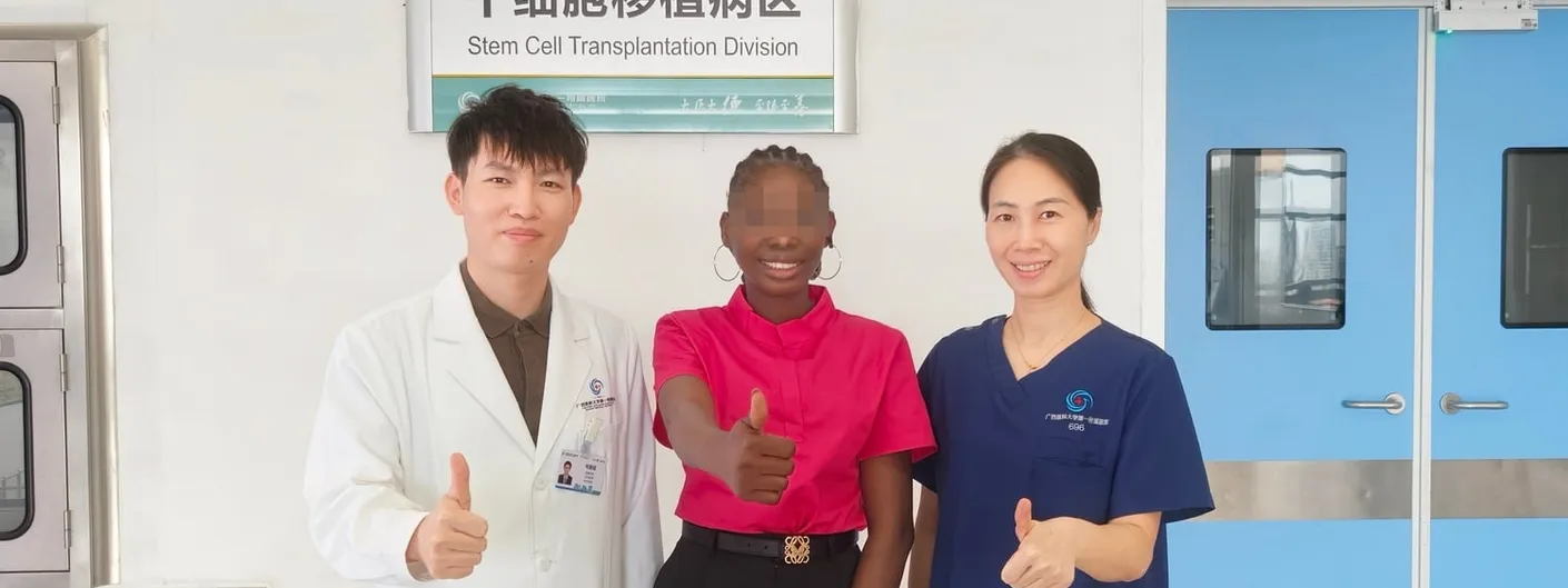 A doctor in a white coat, a patient in a pink shirt whose face is blurred for privacy, and a nurse in navy scrubs stand together giving thumbs up in a hospital corridor beneath a sign reading "Stem Cell Transplantation Division" in Chinese and English.