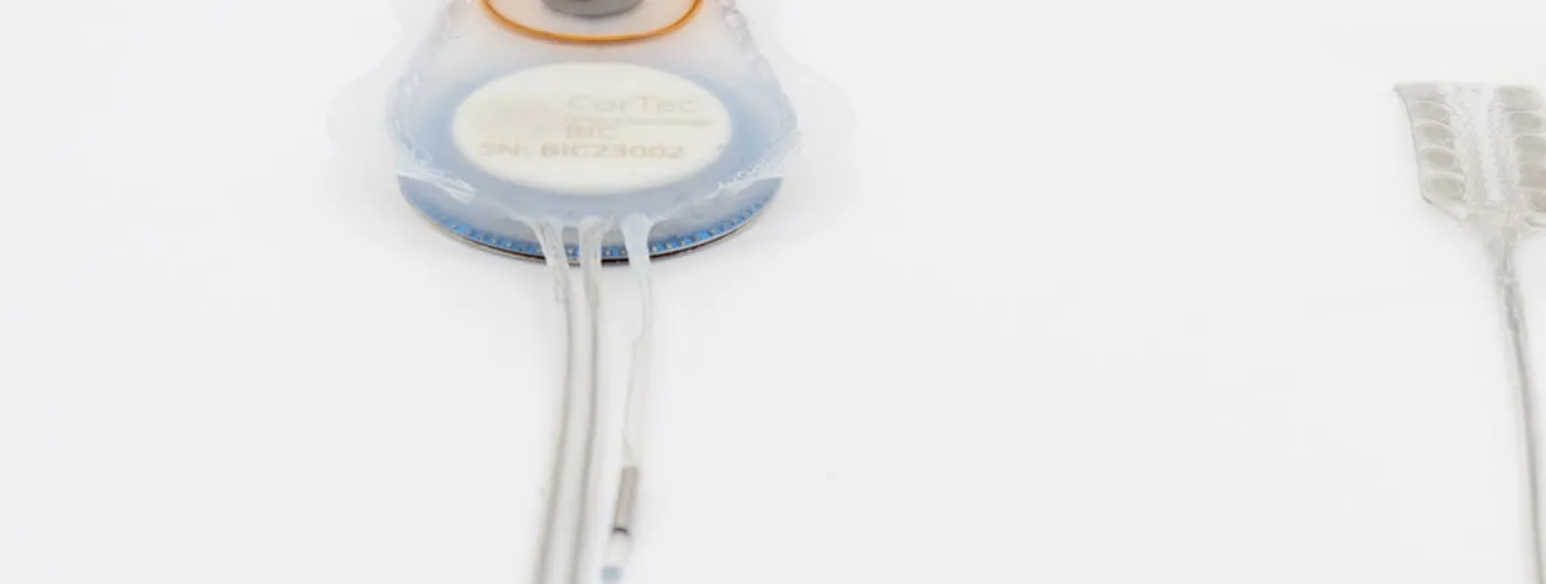 A round implantable device wrapped in translucent mesh, its white centre label reading CorTec Brain Interchange BIC, with three thin lead wires trailing from the lower edge and the end of a flat electrode array visible at the right of the frame.