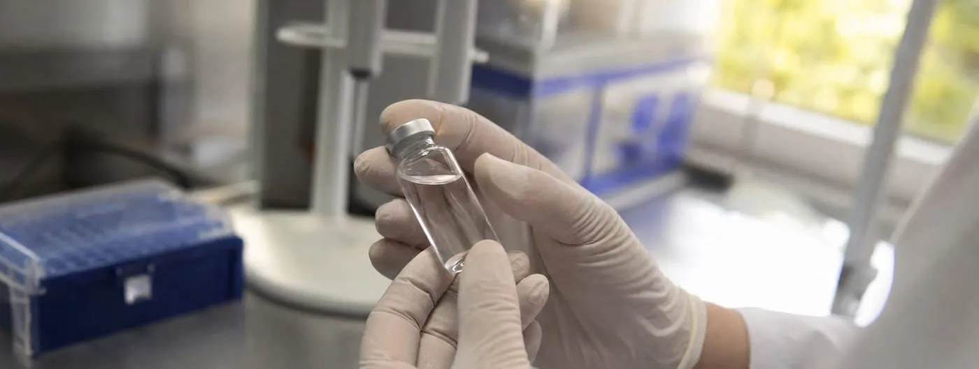 Gloved hands hold a small clear glass vial of colourless solution above a laboratory bench, with a blue pipette-tip box and a bright window out of focus behind.