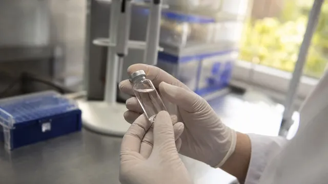 Gloved hands hold a small clear glass vial of colourless solution above a laboratory bench, with a blue pipette-tip box and a bright window out of focus behind.