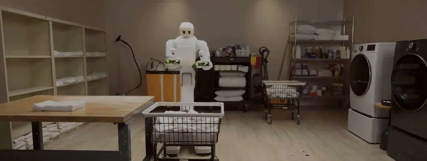A white semi-humanoid robot stands behind a wire laundry cart full of white linens in a laundry room, with a folding table of towels, shelving, a supply cart and front-loading washer and dryer around it.